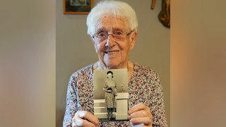 Alte Frau mit weißen Haaren und Brille lächelt in Kamera und hält ein schwarzweiß Foto in den Händen.