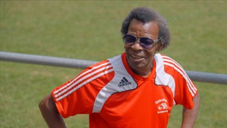 Älterer Mann mit dunkler Haut, Sonnenbrille und rotem Trikot.