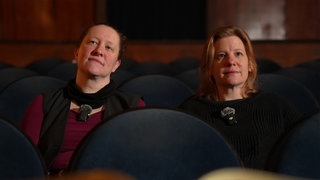 Zwei Frauen sitzen in einem Kino und schauen an der Kamera vorbei nach oben.