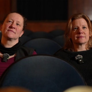Zwei Frauen sitzen in einem Kino und schauen an der Kamera vorbei nach oben.