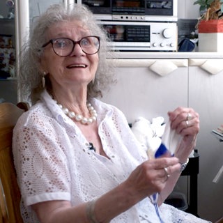 Für ihren Laden hat Gisela 42 Jahre lang gestrickt, gehäkelt und genäht. Sie sitzt in ihrem Wohnzimmer und strickt immer noch weiter. Sie lacht in die Kamera.