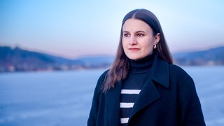 Die ehrenamtliche Sterbebegleiterin Katharina steht vor einem See. Sie trägt ihre Haare offen und hat einen schwarzen Mantel an, unter dem sie einen schwarz-weißen Rollkragenpullover trägt. 