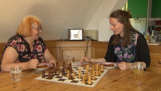 Zwei Frauen sitzen an einem Esstisch und spielen Schach. Barbara, eine Frau um die 60, hat einen weißen Haaransatz, der Rest ist in einem auffälligen Orangene-Rot. Sarah ist in ihren 30ern und hat langes braunes Haar.