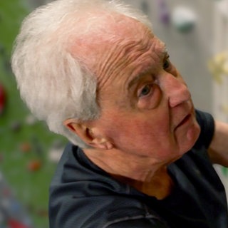 Wilfried, 82 Jahre alt, in einer Kletterhalle. Er ist mit einem Sicherheitsgurt gesichert und hängt an einer Kletterwand. Im Hintergrund sieht man bunte Griffe.
