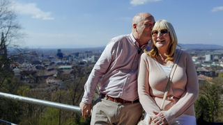 Jutta ist blond und trägt eine große Sonnenbrille. Klaus steht neben ihr und küsst sie auf die Schläfe. Hinter ihnen ist in der Ferne verschwommen Freiburg zu erkennen.