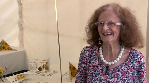 Die 100-jährige Thea feiert ihr Geburtstags-Jubiläum groß und lächelt während sie in die Kamera schaut.