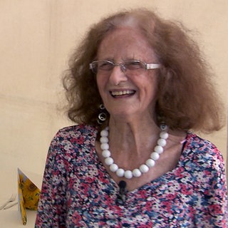 Die 100-jährige Thea feiert ihr Geburtstags-Jubiläum groß und lächelt während sie in die Kamera schaut.