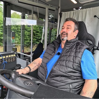 Busfahrer Pit sitzt in einem blauen T-Shirt und schwarzer Pufferweste in seinem Bus hinter dem Lenkrad. Sein Blick ist konzentriert nach vorne gerichtet. Im Hintergrund sieht man Bäume vorbeiziehen.