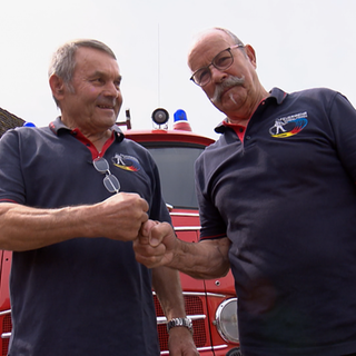 Alfred und Gustav sind Feuerwehrmänner mit grauen Haaren. Gustav trägt einen Schnurrbart. Sie laufen neben einem alten roten Feuerwehrauto.