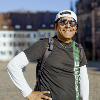 Samuel trägt eine Sonnenbrille und ein weißes Basecap. Er grinst frech in die Kamera und posiert mit einer Hand in der Hüfte aufgestützt.