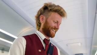 Marius wagt einen Jobwechsel und wird Zugbegleiter. Im Zug führt er Ticketkontrollen durch und trägt eine bordeauxrote Uniform. Marius lächelt dabei.