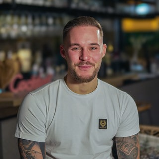 Enrico ist ein junger Mann mit 5-Tage-Bart. Er trägt ein weißes Shirt und hat tätowierte Arme.