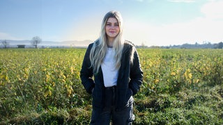 Vanessa steht ganz allein auf einem Feld. Im Hintergrund sind grüne Felder zu sehen. Sie trägt eine schwarze Jacke und hat lange blonde Haare. Sie schaut in die Kamera. 