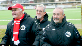 Drei Männer im Rentenalter, die sich im Fußballverein ehrenamtlich engagieren,  stehen am Spielfeldrand und tragen Fan-Artikel des SC Bahlingen.