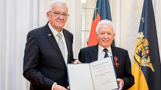 Für sein besonderes ehrenamtliches Engagement bekommt Helmut das Bundesverdienstkreuz verliehen. Auf dem Foto hält Helmut die Urkunde dazu in der Hand, neben ihm steht der baden-württembergische Ministerpräsident Winfried Kretschmann. Dieser übergibt Helmut die Urkunde. Hinter den beiden steht eine Deutschlandflagge und die Flagge von Baden-Württemberg. 