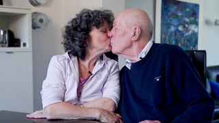 Ehepaar Julia und Gunar sitzen bei sich zu Hause am Esstisch und küssen sich.