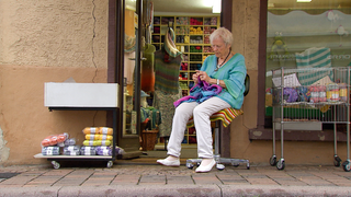 95 Jahre alte Frau sitzt vor dem Eingang ihres Wollgeschäfts auf einem Drehstuhl und strickt. Sie trägt einen gestrickten, blau-grünen Pullover und eine weiße Hose und weiße Ballerinas. Sie schaut auf das lila Strickzeug auf ihrem Schoß.