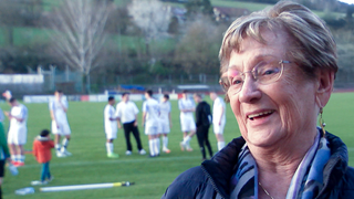 Waltraud steht am Rand eines Fußballplatzes und lacht. Auf dem Fußballfeld hinter ihr sind Fußballspieler zu sehen.