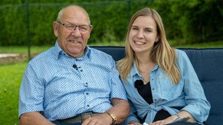 Ein älterer Herr und eine junge Frau sitzen im Freien auf einer gepolsterten Bank. Er trägt ein blaues Hemd und sie eine blaue Bluse.