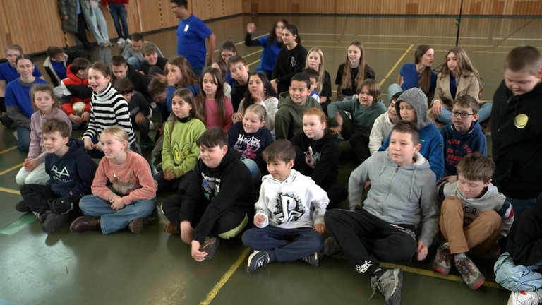 Grundschulkinder sitzen auf dem Boden einer Turnhalle