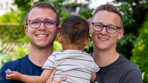 Zwei Männer halten ein Kleinkind auf dem Arm und lachen in die Kamera. Jonas und Jan haben viele Hürden genommen, um ein Kind adoptieren zu können.