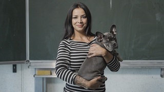 Eine Frau mit schwarzen Haaren hält eine französische Bulldogge auf dem Arm. Lehrerin Julia paukt mit ihren Schülern dreimal die Woche für den Hauptschulabschluss.