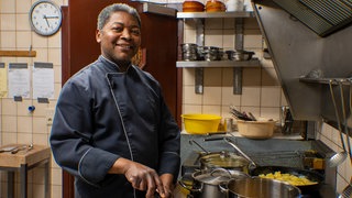 Schwarzer Mann in blauer Kochkleidung steht an Herd.