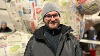 Junger Mann, mit Brille und grauer Mütze lächelt vor Fastnachtswagen in die Kamera.
