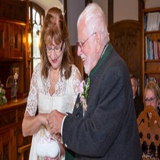 Edith und Harald steigen bei ihrer Hochzeit durch ein großes Herz. Sie trägt ein weißes Dirndl und er eine braune Lederhose. 