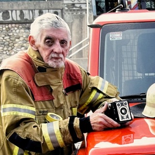 Bertold ist Feuerwehr-Fotograf. Er trägt Feuerwehrbekleidung und hat seine Kamera in der Hand. Er steht neben einem Einsatzwagen und schaut in die Kamera.