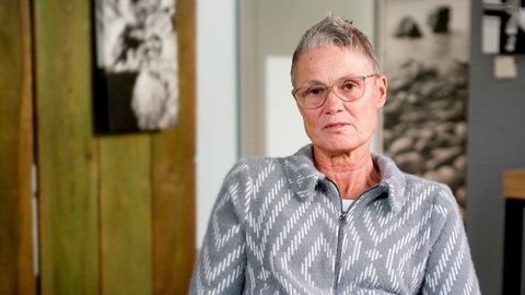 Sabine, Tochter eines Opfers des RAF-Anschlags von 1977 in Karlsruhe, sitzt in einem Raum und spricht über den Verlust ihres Vaters. Sie trägt ein grau gemustertes Oberteil und eine Brille.