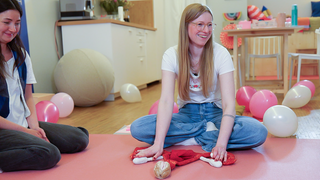 Julia sitzt auf dem Boden, im Schneidersitz, vor ihr liegt ein Plüsch-Modell eines Uterus und sie lächelt. Julia ist gerade dabei, einen Menstruationskurs zu geben, um sie herum liegen Luftballons und auch eine Teilnehmerin ist zu sehen.