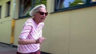 Magdalene ist 95 Jahre alt. Sie trägt eine Sonnenbrille und rosa Oberteil. Magdalene ist auf einem Sportplatz und ist gerade losgerannt. Ihre grauen, schulterlangen Haare wehen nach hinten.