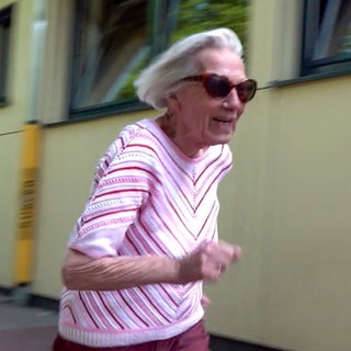 Magdalene ist 95 Jahre alt. Sie trägt eine Sonnenbrille und rosa Oberteil. Magdalene ist auf einem Sportplatz und ist gerade losgerannt. Ihre grauen, schulterlangen Haare wehen nach hinten.