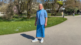 Gerd ist 70. Gerd trägt langes graues Haar in einem Pferdeschwanz und ein fliederfarbenes Kostüm. Gerd steht in der Lichtentaler Allee in Baden-Baden und lächelt in die Kamera. 