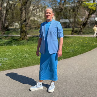 Gerd ist 70. Gerd trägt langes graues Haar in einem Pferdeschwanz und ein fliederfarbenes Kostüm. Gerd steht in der Lichtentaler Allee in Baden-Baden und lächelt in die Kamera. 