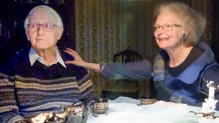 Das Ehepaar Dieter und Angelika aus Wildberg sitzen gemeinsam am Esstisch. Er trägt einen gestreiften Stickpullover und ein Hemd darunter, Angelika einen blauen Pullover und eine Weste darüber. Sie streckt ihren Arm aus und berührt die Schulter ihres 19 Jahre älteren Partners. 