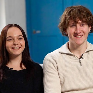 Die Freunde Melina und Dante sitzen auf einem Sofa vor einer blauen Wand. Melina hat dunkle, schulterlange Haare, trägt einen blauen Pullover und Jeans. Dante sitzt rechts neben ihr. Er ist etwas größer als Melina, hat hellbraune Haare und trägt einen weißen Pullover und braune Hosen. Sie sind schon seit ihrer Kindheit befreundet und ihre Freundschaft hat auch Melinas Krebsdiagnose und – therapie überdauert.