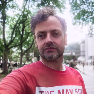 Andreas macht bei einem Halbmarathon ein Selfie von sich. Er steht auf einer breiten Straße, rechts und links davon stehen viele Bäume, im Hintergrund sind noch andere Menschen in Sportklamotten zu sehen. Er trägt ein rotes T-Shirt mit der Aufschrift: The May 50k.