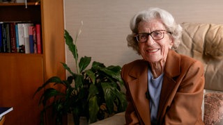 Die 104-jährige Waltraud aus Pforzheim sitzt lächelnd in ihrem Wohnzimmer. Die Seniorin trägt eine braune Jacke und eine Brille, im Hintergrund stehen ein Bücherregal und eine Zimmerpflanze. Die Seniorin engagiert sich seit Jahrzehnten für den Wildpark Pforzheim und verrät ihr Geheimnis fürs Altwerden im Interview. 