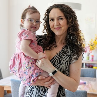 Laura hält ihre 2-jährige Tochter Elisa auf dem Arm. Elisa hat das Angelman-Syndrom und trägt eine Brille, Laura hat braune Locken und trägt ein Sommerkleid. Beide lachen in die Kamera.