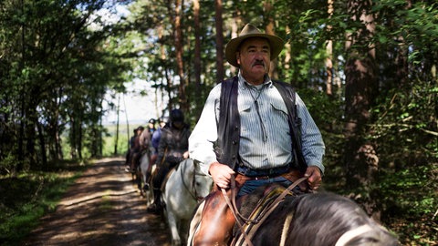 Mann mit Cowboyhut und Schnurrbart sitzt auf Pferd, hinter ihm andere Reiter.