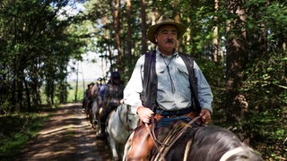 Mann mit Cowboyhut und Schnurrbart sitzt auf Pferd, hinter ihm andere Reiter.