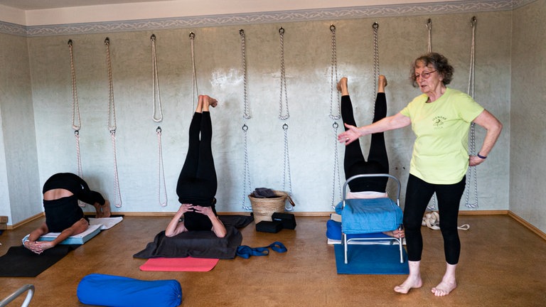 Frauen üben Yoga, Übungsleiterin steht vorne im grünen T-Shirt und leitet die Frauen an.