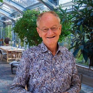 Mittelalter Mann in buntem Hemd, der vor seinem Lehmhaus steht, das in ein Glashausgebaut ist.