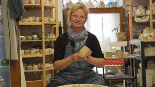 50-Jährige vor einer Töpferscheibe mit Tongefäß in den Händen
