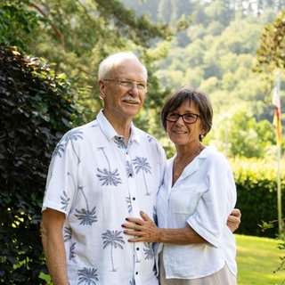 Altes Paar im Garten, das sich in den Armen hält. Beide tragen ein weißes Oberteil.
