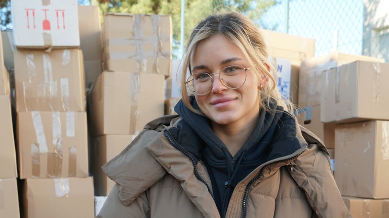 Junge Frau mit dicker Jacke, schwarzem Kapuzenpullover und Brille vor etlichen Kartons.