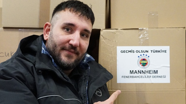 Mann vor einer Paketwand. Auf einem Paket klebt ein Zettel mit der Aufschrift: ‘Gute Besserung, Türkei!’