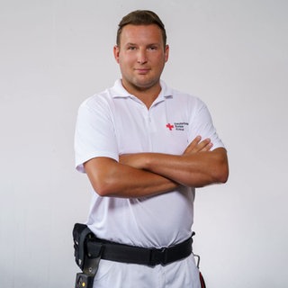 Notfallsanitäter Chris steht in seiner Rettungsdienst-Uniform vor einem grauen Hintergrund und lächelt in die Kamera.  